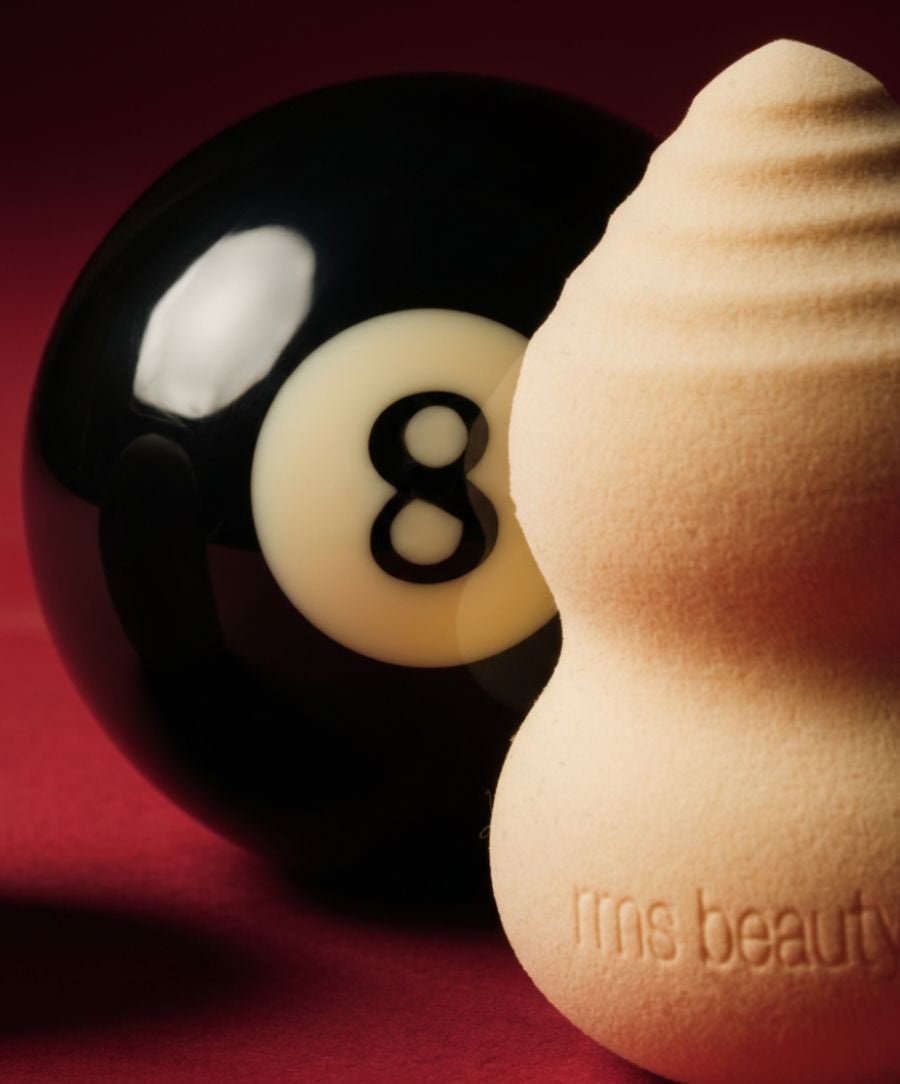 Close-up of the Skin2Skin Beauty Sponge made of soft material embossed with rms beauty logo, positioned next to a black and white billiard 8 ball on a red surface.