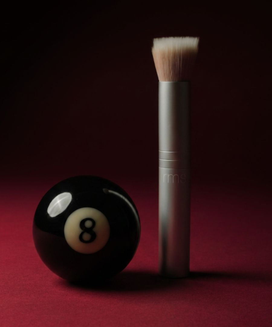 Skin2Skin Blush Brush standing upright next to a black eight ball on a deep red surface, highlighting the brush’s dense, angled bristles and sleek silver handle.