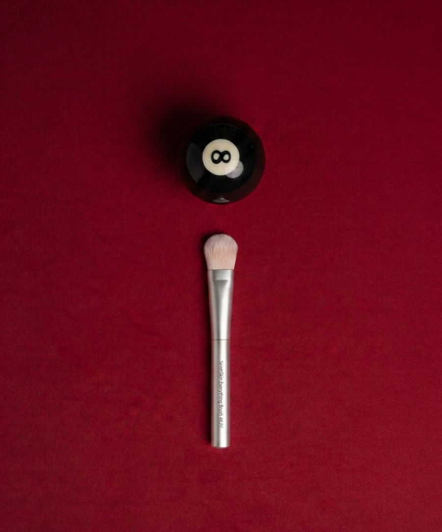 Skin2Skin Everything Brush with soft bristles and silver handle placed on a deep red background next to a black and white magic 8 ball.