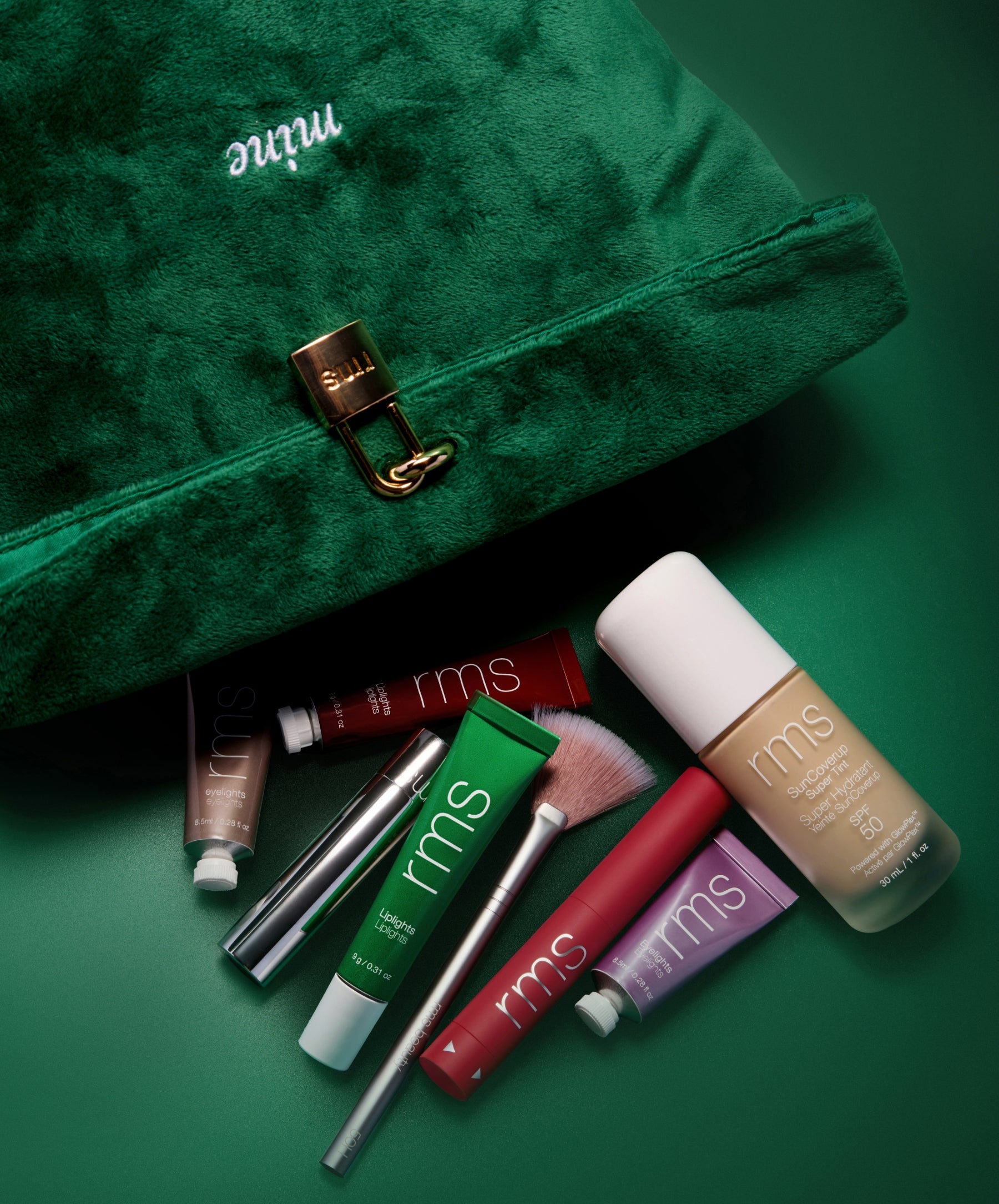 The Emerald Luxe Pouch in rich green velvet with gold clasp, surrounded by RMS Beauty skincare and makeup products including foundation, lip tints, and brushes on a matching green background.