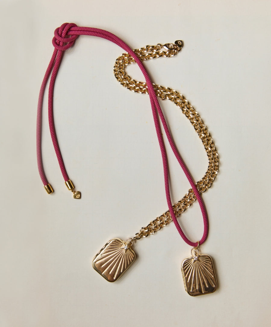 Two gold-tone lockets with sunburst designs, one on a pink cord necklace and the other on a gold chain, styled on a light background featuring the HART x RMS: Lip2Cheek Locket.