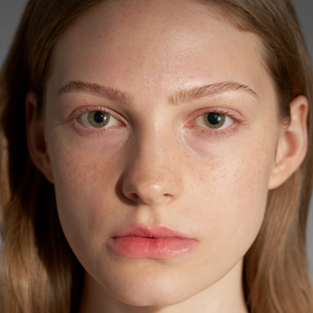 Close-up of a young woman with clear, natural skin, light freckles, soft pink lips, and light brown hair against a neutral background.