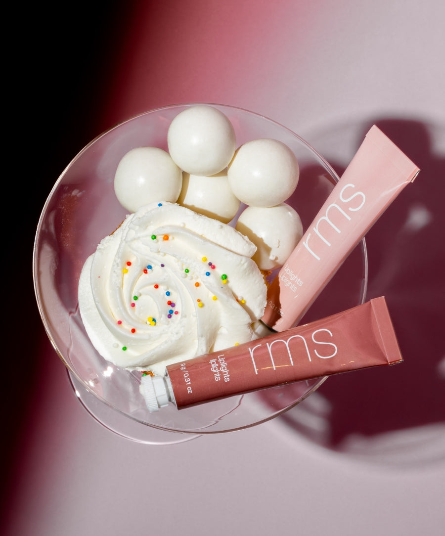 Two tubes of Liplights Cream Lip Gloss displayed in a glass bowl with white gumballs and a swirl of whipped cream topped with colorful sprinkles, highlighting a clean beauty and natural ingredients theme.