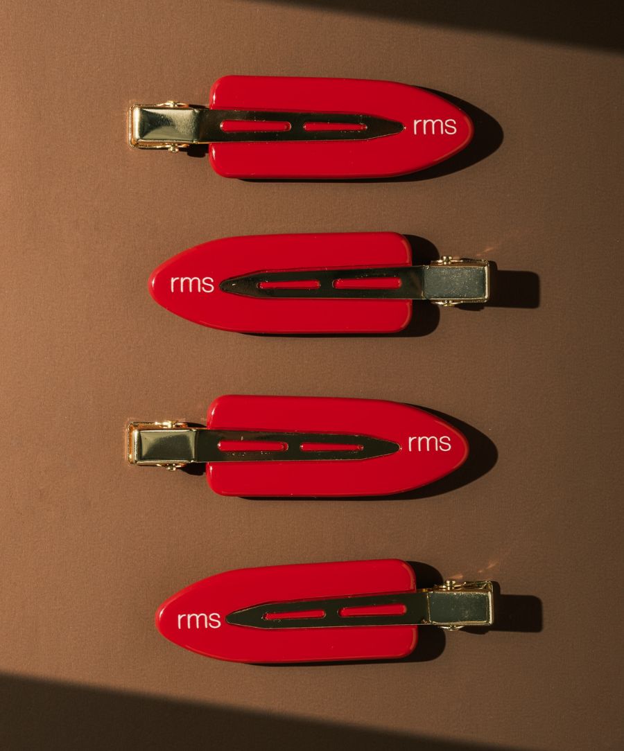 Four RMS Creaseless Red Hair Clips arranged in a vertical line on a brown surface, featuring a vibrant red color with gold-tone metal accents and the "rms" logo on each clip.