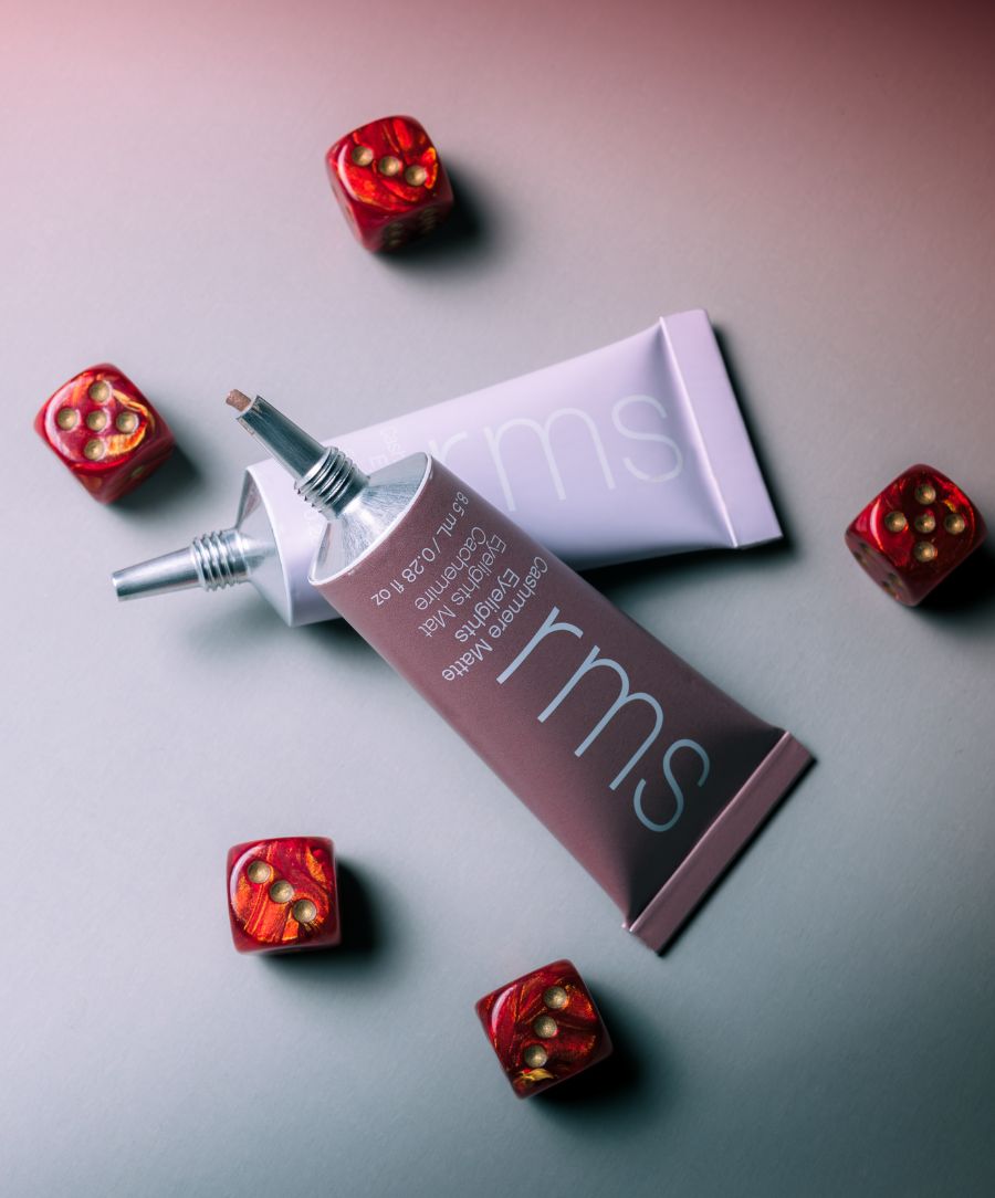 Two tubes of Cashmere Matte Eyelights makeup in soft lavender packaging surrounded by small red dice with gold dots on a gradient background.