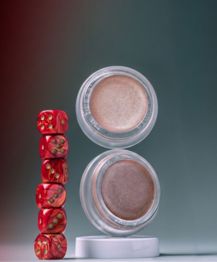 Two open jars of Luminizer cream makeup with a stack of red dice beside them on a smooth surface against a muted dark background