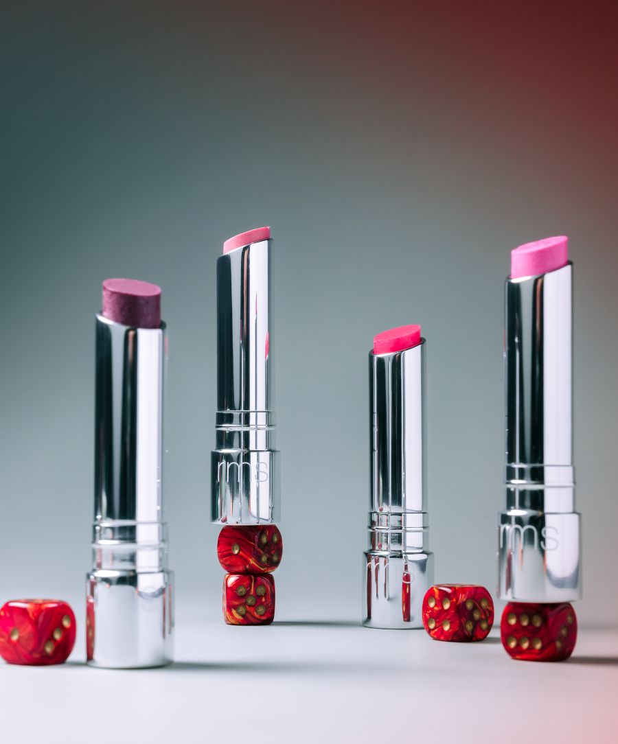 Four tubes of Tinted Daily Lip Balm in varying pink and red shades are displayed standing upright and balanced on small red dice with gold dots, against a gradient background.