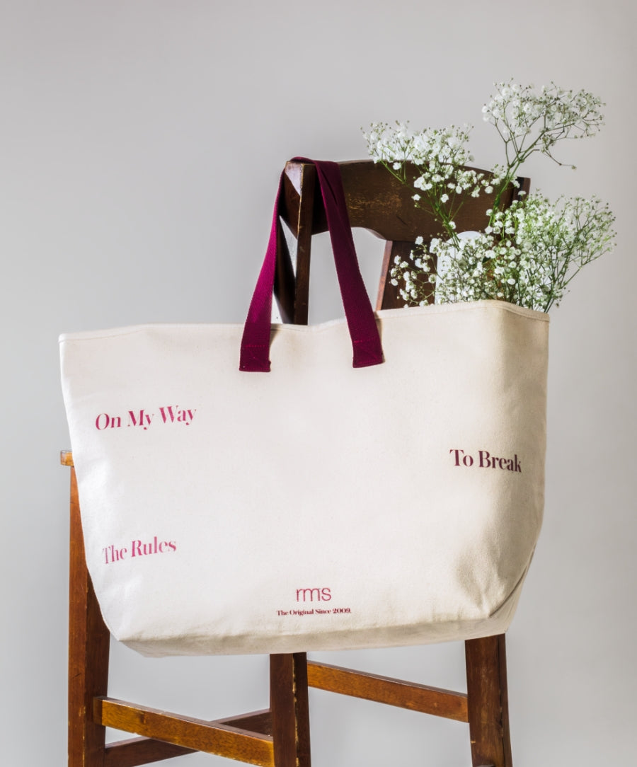 The Weekender Travel Tote in natural fabric with dark red handles, featuring printed text and filled with delicate white flowers, resting on a wooden chair against a plain background.