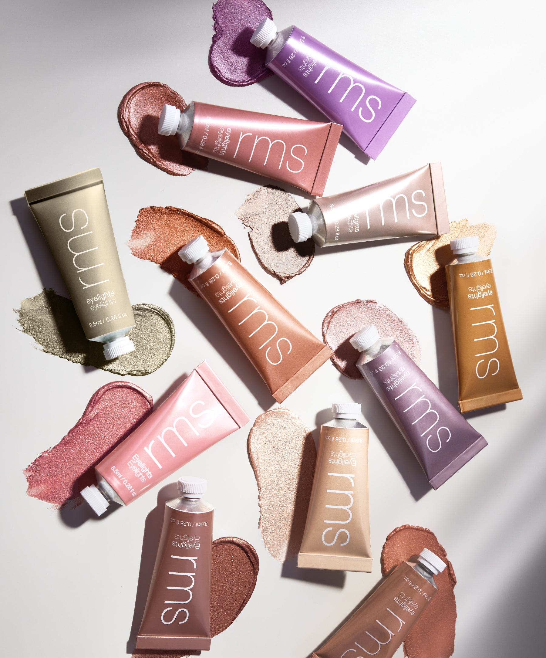 Various colorful tubes of Eyelights Cream Eyeshadow from RMS Beauty arranged with swatches of different shades on a white surface, showcasing creamy texture and clean beauty makeup.