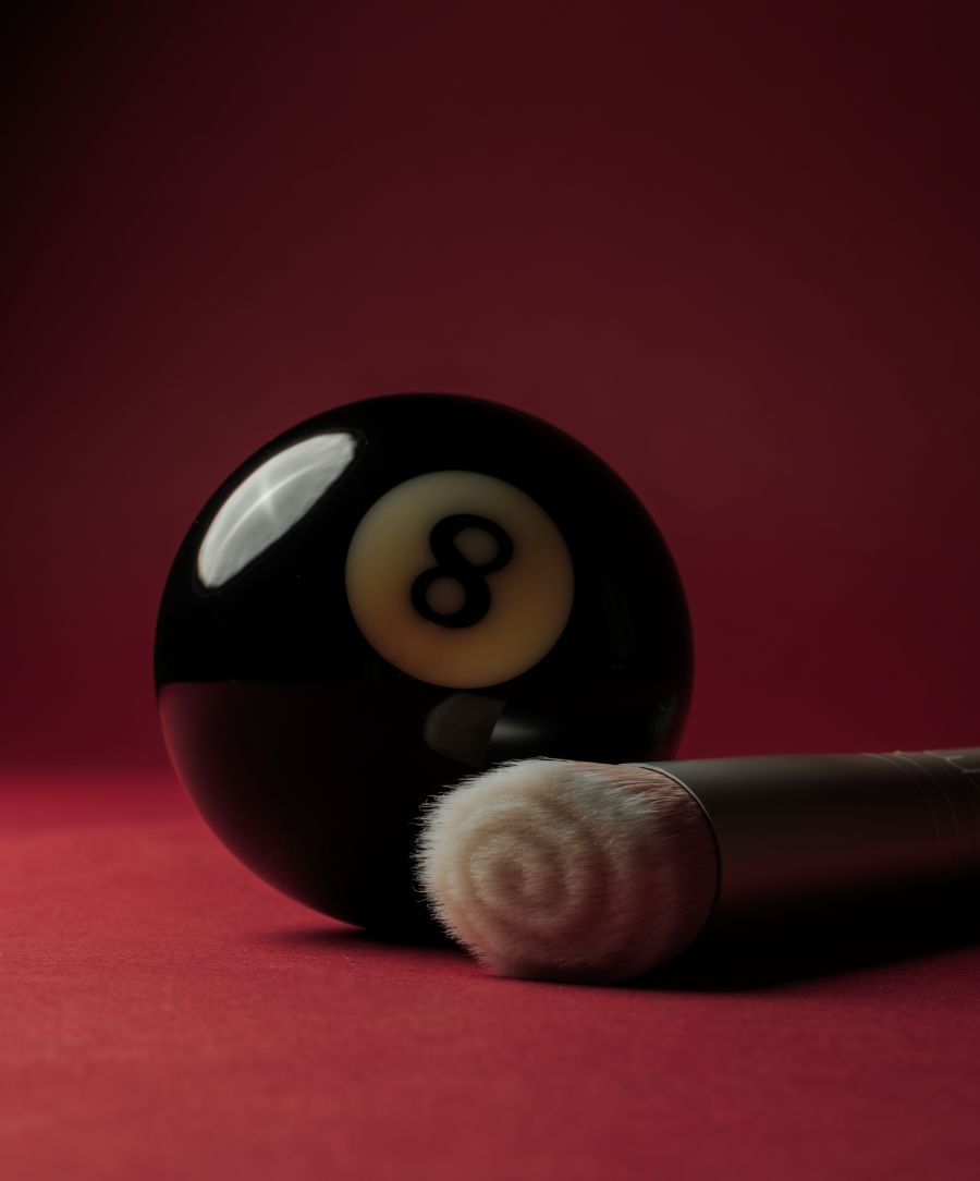 Skin2Skin Foundation Brush with soft, densely packed bristles next to a black eight ball on a deep red background, highlighting its smooth texture for flawless makeup application.