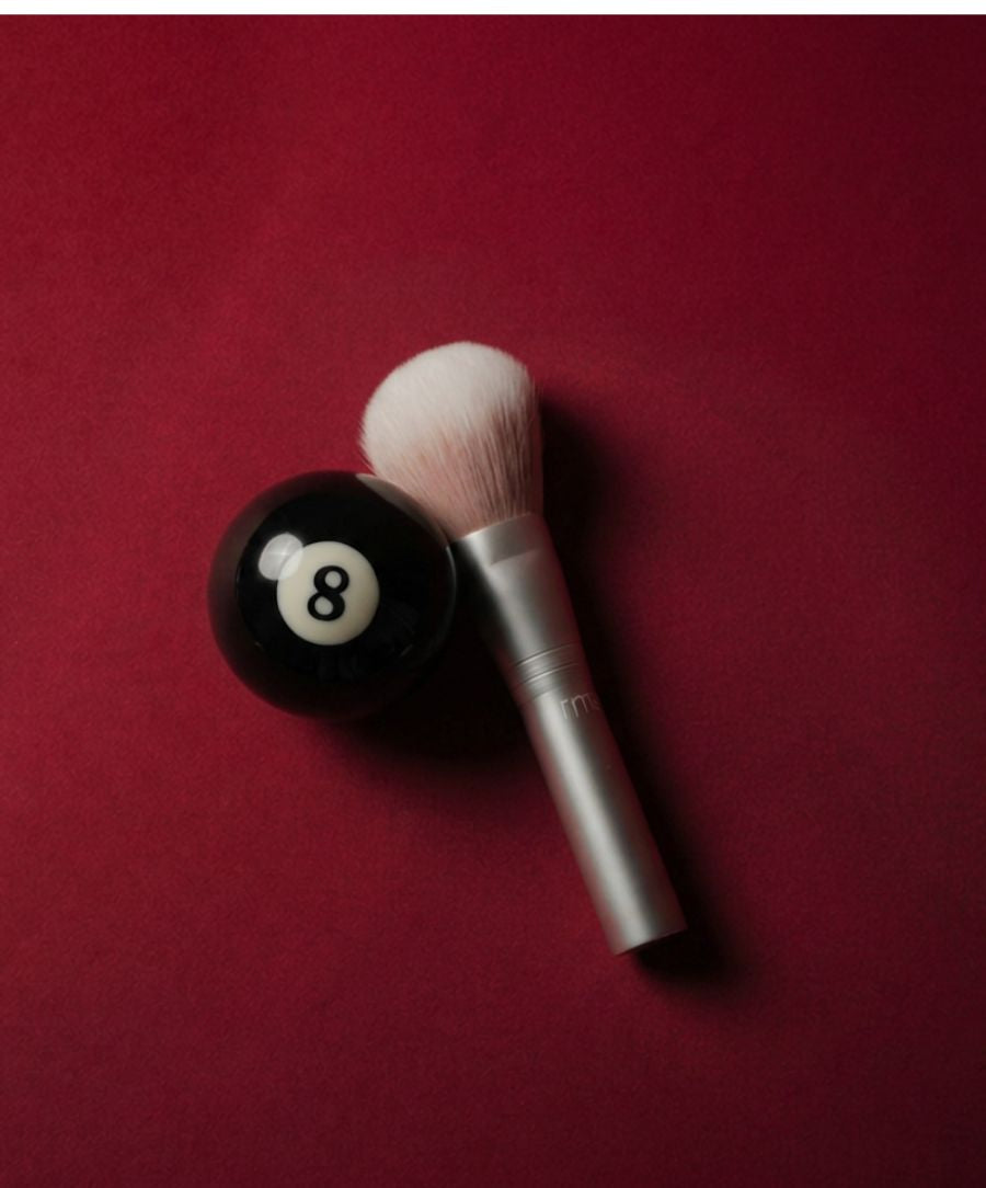 Skin2Skin Powder Blush Brush with soft white bristles and silver handle placed next to a black eight ball on a deep red background, highlighting clean beauty and cruelty free makeup.