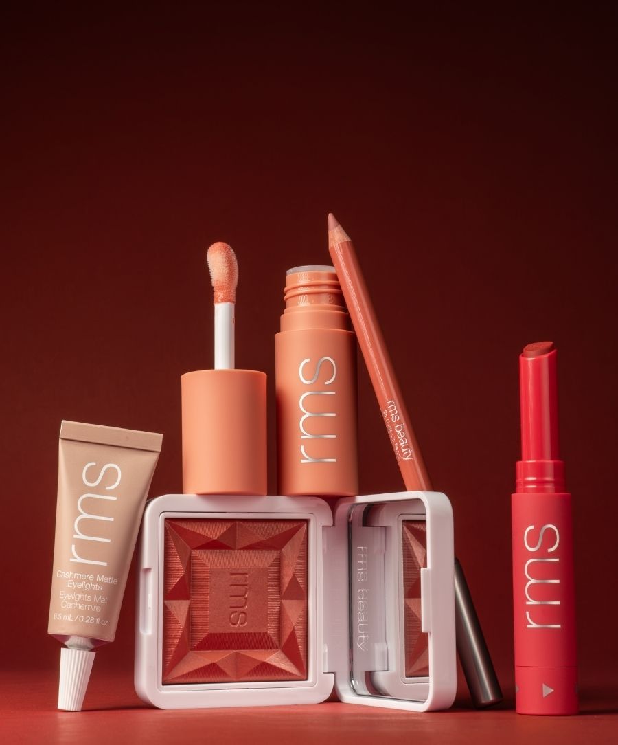 An arrangement of RMS Beauty makeup products in warm, sun-kissed tones, featuring a lip balm, lip gloss, lip liner, cream eyeshadow, and a blush from the Color Capsule: The Summer Sangria Edit collection, highlighting clean ingredients and sustainable packaging.