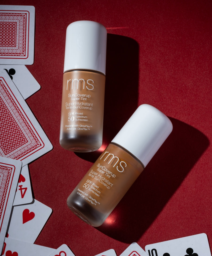 Two bottles of SunCoverup Super Tint Broad Spectrum SPF 50 Sunscreen by RMS Beauty placed on a red surface with scattered playing cards, highlighting clean beauty and sustainable packaging.