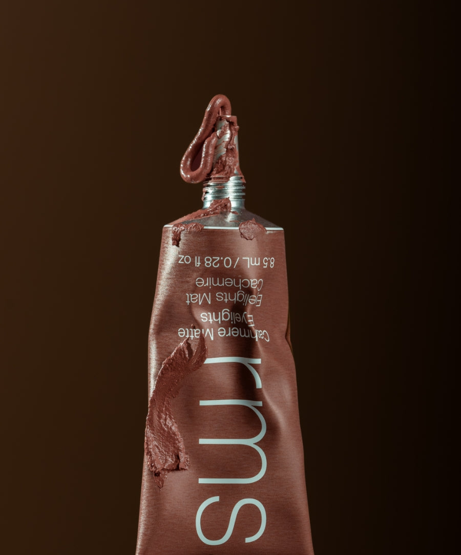 Close-up of a partially squeezed tube of Cashmere Matte Eyelights makeup in a warm brown shade, showcasing smooth creamy texture with matte finish against a dark brown background.