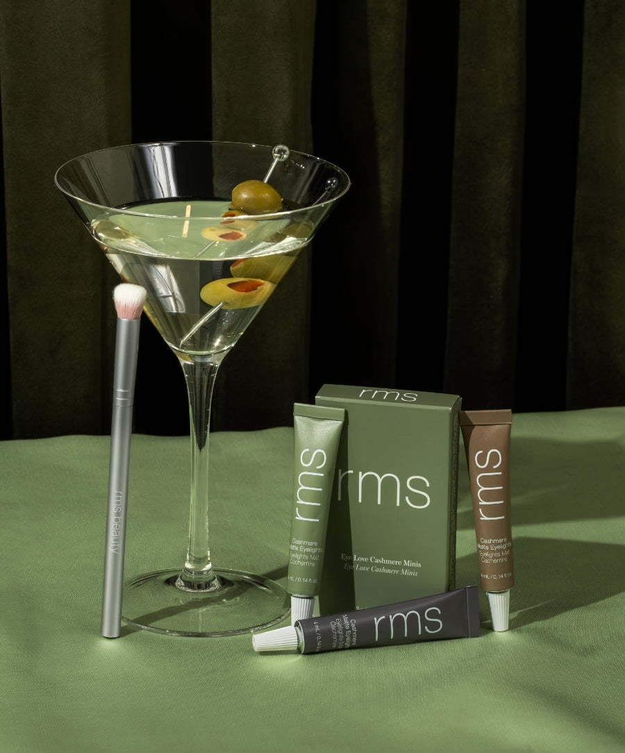 Minimalist arrangement featuring the Mini Cashmere Eyelights Trio + Eye Polish Brush with three tinted tubes and a soft brush next to a martini glass garnished with olives.