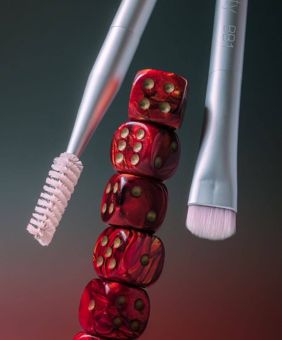 Close-up of two silver Back2Brow Brush tools positioned near a stack of glossy red dice, highlighting the textured bristle and angled brush head for precise eyebrow grooming.