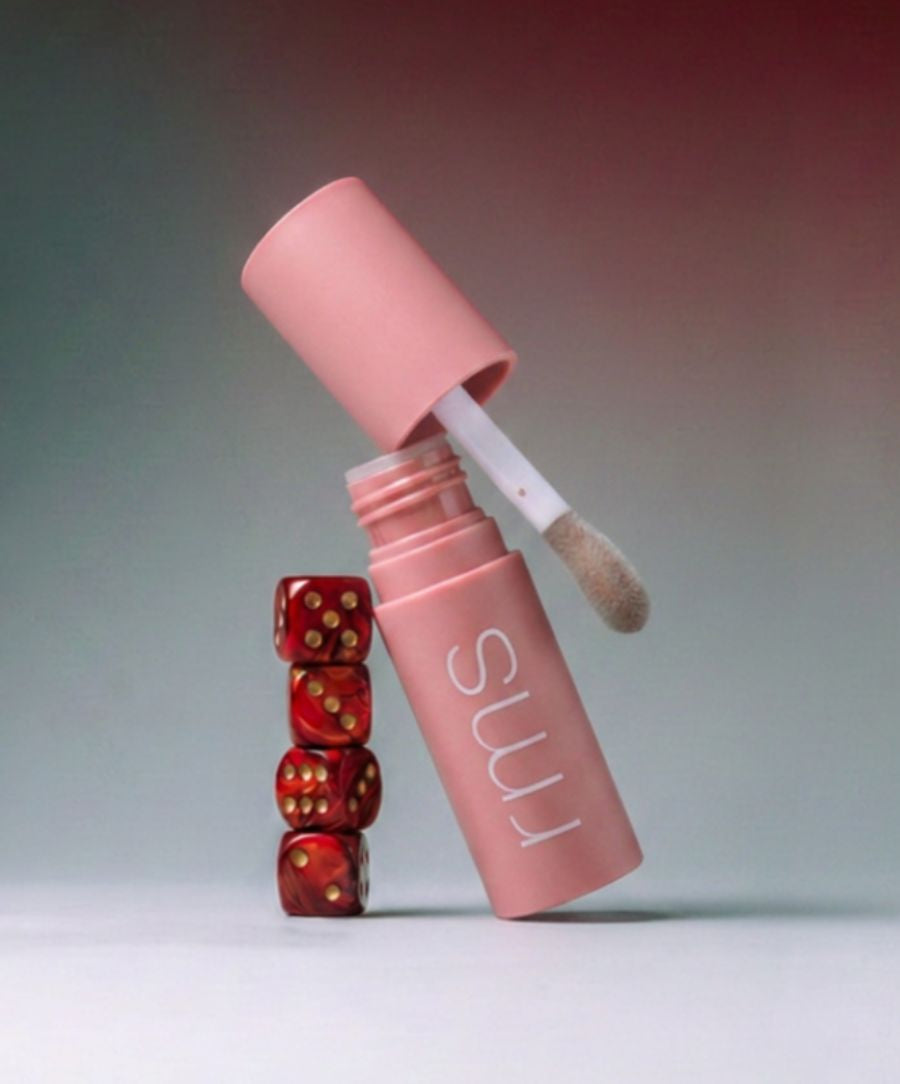 A pink tube of Legendary Lip Oil with a white applicator wand resting on top of a stack of four red dice, highlighting clean beauty and natural skincare infused makeup.