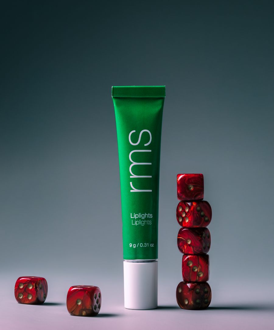 Liplights Cream Lip Gloss in a green tube displayed with stacked and scattered red dice, highlighting a clean beauty, cruelty free makeup product with natural and organic ingredients.