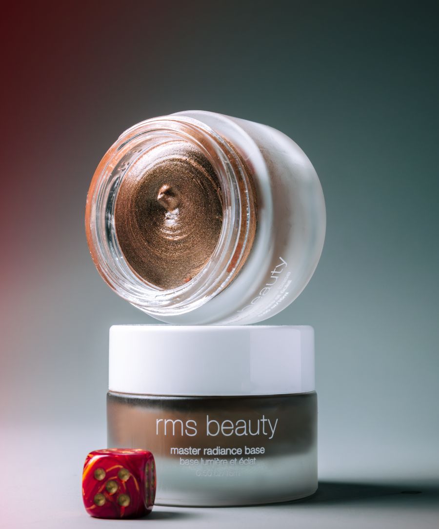 Open jar of Master Radiance Base by RMS Beauty showing the creamy bronze shimmer texture on top of the closed frosted glass container with white lid.