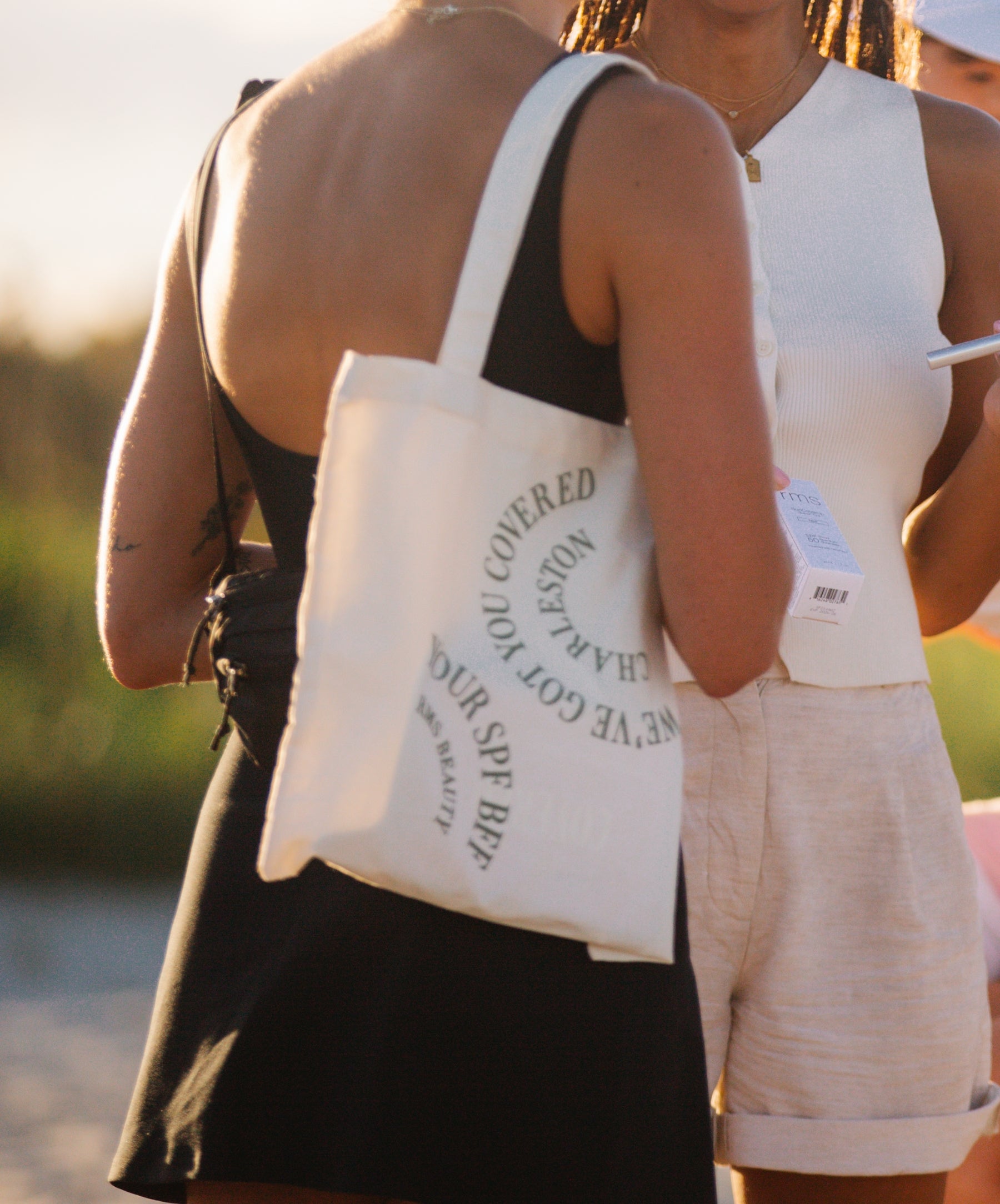 Woman wearing a black dress carrying a white canvas tote bag with circular text design, featuring the We've Got You Covered Charleston Tote in a sunny outdoor setting.