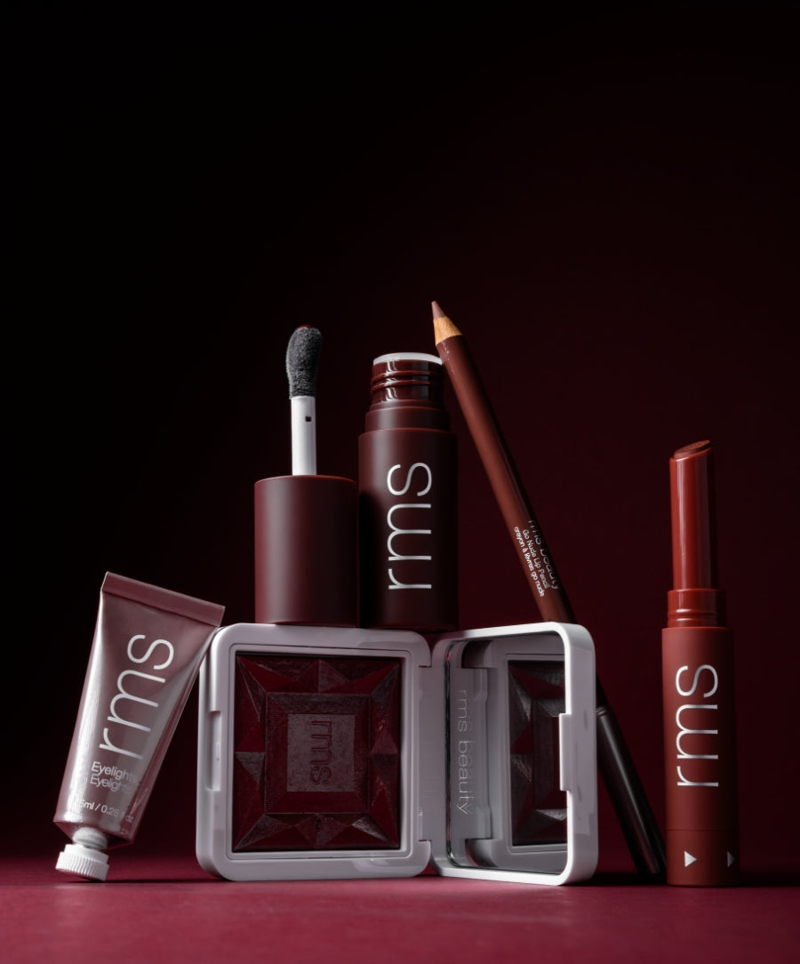 A collection of RMS Beauty makeup products in deep wine tones including a lip gloss, pencil, cream blush, lipstick, and eye shadow with a dark red background, featuring Color Capsule: The Wine Edit.