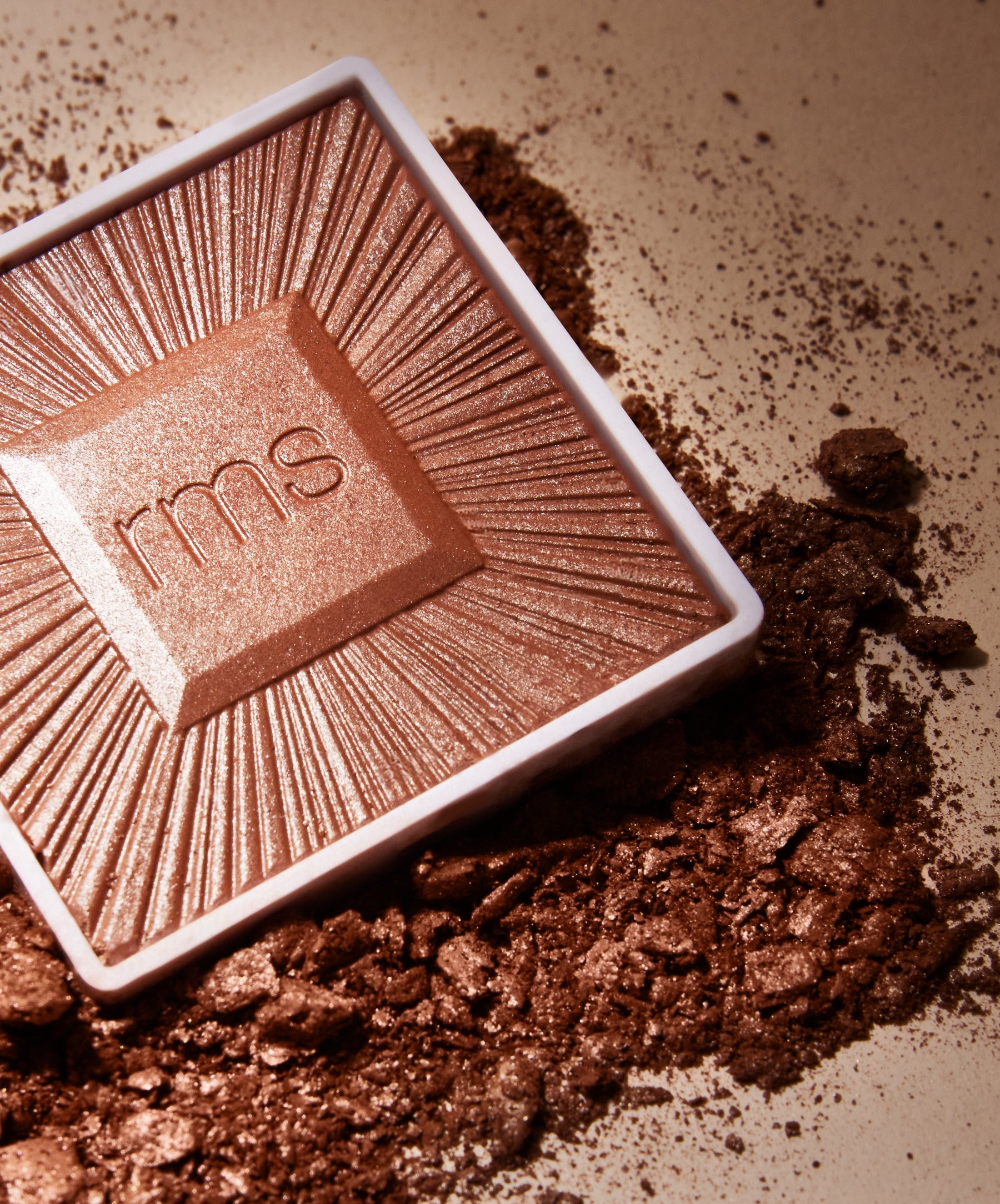 Close-up of a shimmering bronze powder makeup embossed with rms, surrounded by crushed pigment, showcasing the ReDimension Hydra Bronzer with a radiant finish.