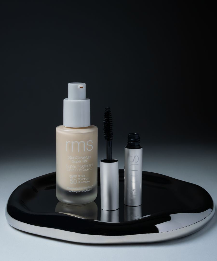 SunCoverup Super Tint Broad Spectrum SPF 50 Sunscreen in a pump bottle beside an open mascara tube, both displayed on a reflective black irregular tray against a dark background.