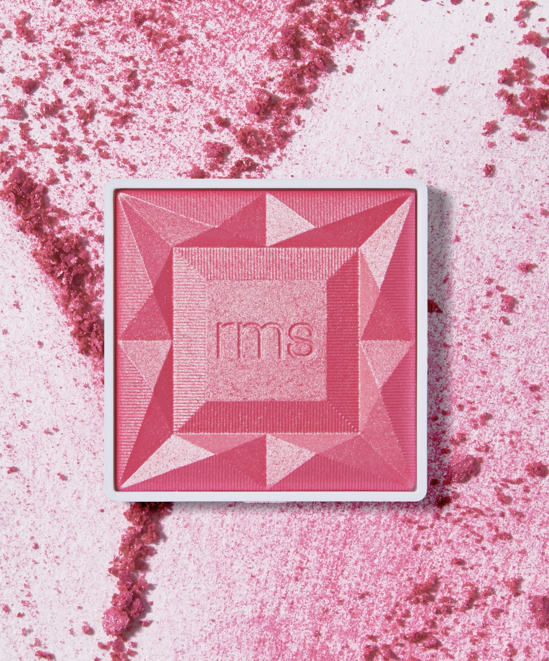 Square compact of ReDimension Hydra Powder Blush in a vibrant pink shade with geometric embossed design and RMS branding, surrounded by crushed powder on a light background.