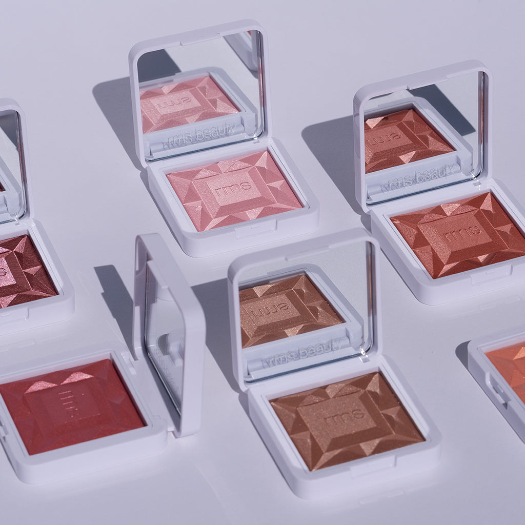 Open compact cases of ReDimension Hydra Powder Blush in various warm and neutral shades displayed on a light surface with natural lighting highlighting their textured powder and clean beauty appeal.