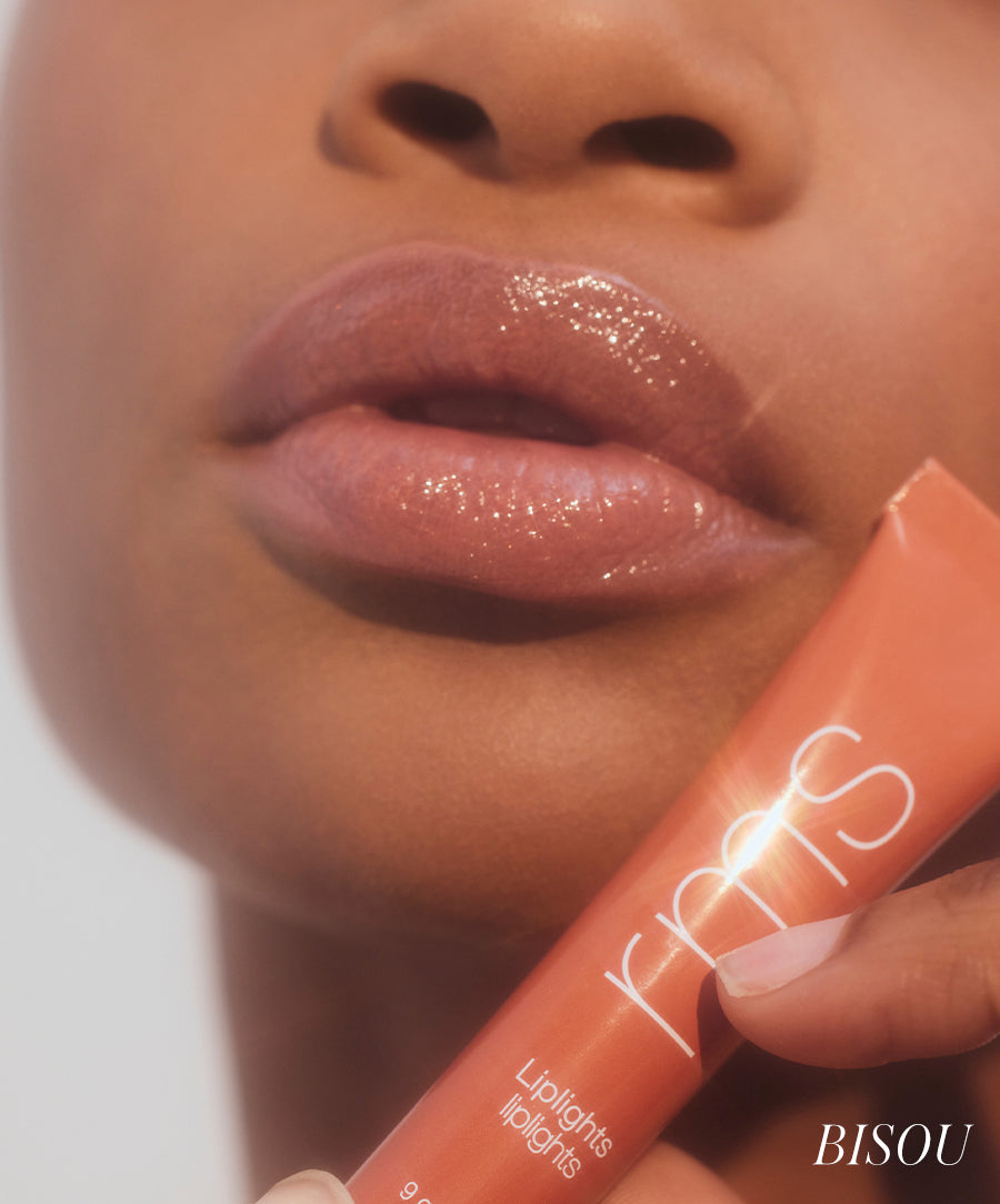 Close-up of a person with smooth skin wearing glossy lips holding a tube of Liplights Cream Lip Gloss by RMS Beauty, showcasing clean beauty and natural ingredients.